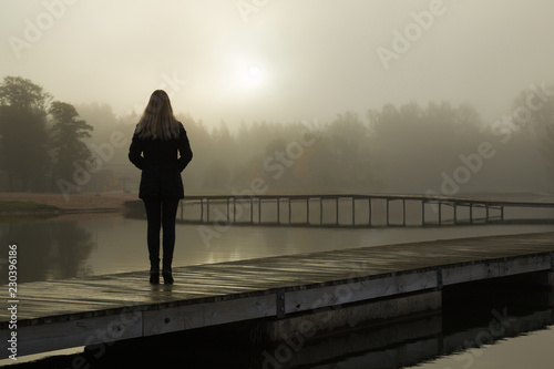 Young woman standing alone on lake footbridge and staring at sunrise in gray, cloudy sky. Mist over water. Foggy air. Early chilly morning. Dark, scary moment and gloomy atmosphere. Back view.