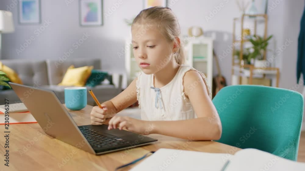 Smart Little Girl Does Homework in Her Living Room. She's Sitting at Her Desk, Uses Laptop and Writes with a Pen in Her Textbooks. Smart Little Girl Does Homework in Her Living Room. She's Sitting at Her Desk, Uses Laptop and Writes with a Pen in Her Textbooks.