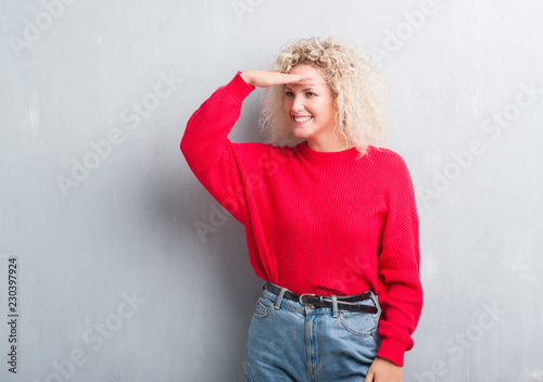 Young blonde woman with curly hair over grunge grey background very happy and smiling looking far away with hand over head. Searching concept.