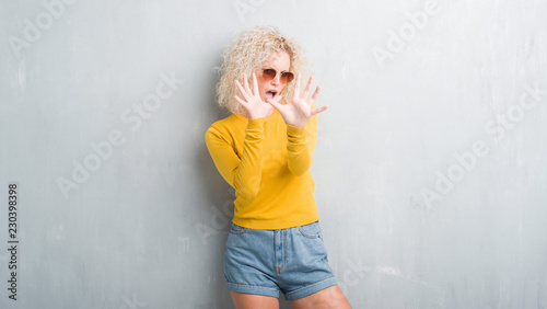 Young blonde woman with curly hair over grunge grey background afraid and terrified with fear expression stop gesture with hands, shouting in shock. Panic concept.