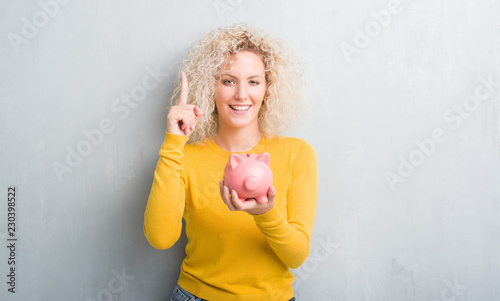 Young blonde woman over grunge grey background holding piggy bank surprised with an idea or question pointing finger with happy face, number one
