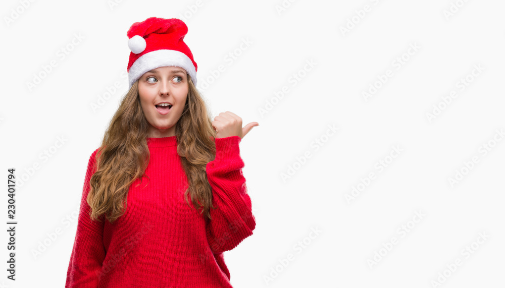 Young blonde woman wearing santa claus hat pointing and showing with thumb up to the side with happy face smiling Young blonde woman wearing santa claus hat pointing and showing with thumb up to the side with happy face smiling