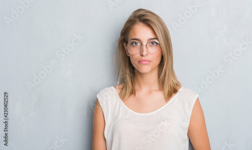 Beautiful young woman standing over grunge grey wall wearing glasses with a confident expression on smart face thinking serious