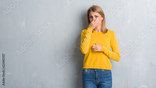Beautiful young woman standing over grunge grey wall smelling something stinky and disgusting, intolerable smell, holding breath with fingers on nose. Bad smells concept.