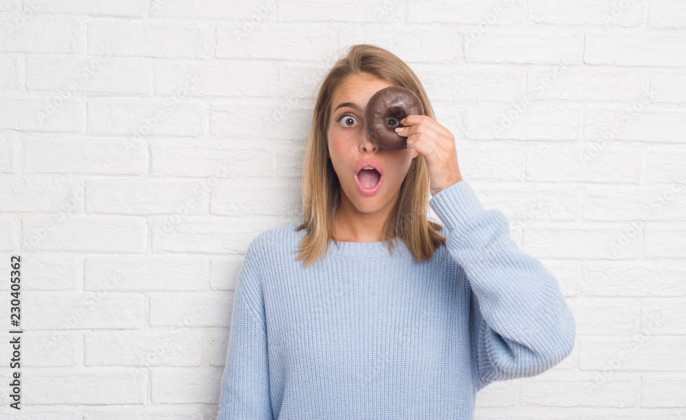 Beautiful young woman over white brick wall eating chocolate donut scared in shock with a surprise face, afraid and excited with fear expression Beautiful young woman over white brick wall eating chocolate donut scared in shock with a surprise face, afraid and excited with fear expression