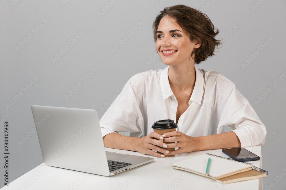Happy cheerful business woman posing isolated over grey wall background sitting at the table using laptop. Happy cheerful business woman posing isolated over grey wall background sitting at the table using laptop.