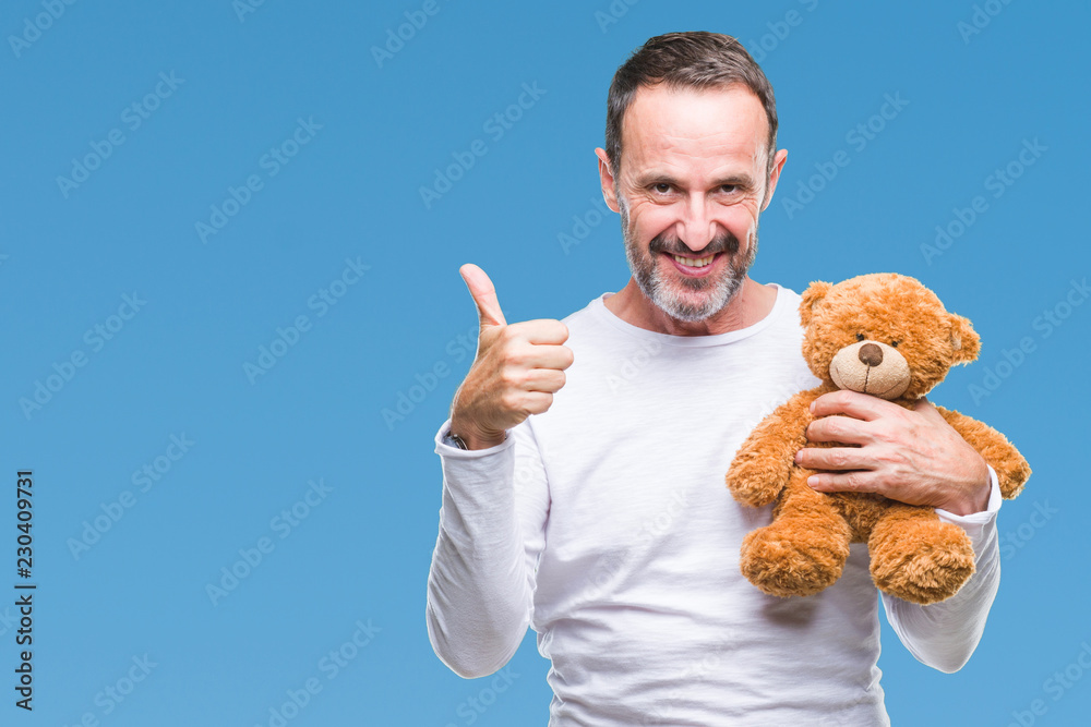 Middle age senior hoary man holding teddy bear over isolated background happy with big smile doing ok sign, thumb up with fingers, excellent sign Middle age senior hoary man holding teddy bear over isolated background happy with big smile doing ok sign, thumb up with fingers, excellent sign