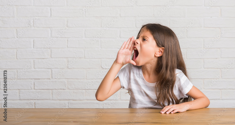 Young hispanic kid sitting on the table at home shouting and screaming loud to side with hand on mouth. Communication concept. Young hispanic kid sitting on the table at home shouting and screaming loud to side with hand on mouth. Communication concept.