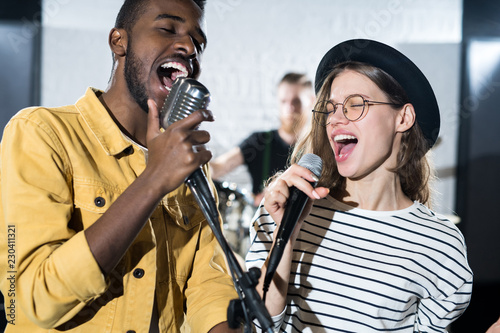 Waist up portrait of two young people enjoying singing with microphones during band performance on stage
