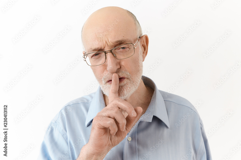Strict annoyed elderly chief executive officer in blue shirt and spectacles making shushing sign during conference, asking to speak quietly. Senior man holding fore finger at lips, saying Shh Strict annoyed elderly chief executive officer in blue shirt and spectacles making shushing sign during conference, asking to speak quietly. Senior man holding fore finger at lips, saying Shh