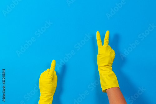 Woman hands with yellow rubber gloves making a gesture meaning victory and points upwards with finger, blue background