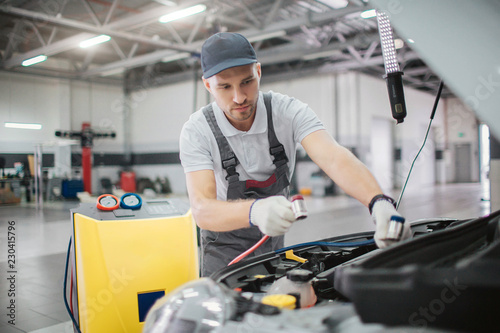 Worker stands at opened front part of car body. He holds power cords for connecting it to engine.