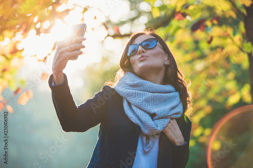 Beautiful young woman making selfie with smartphone in a park. Autumn romantic mood. Real sun flare on sunset.