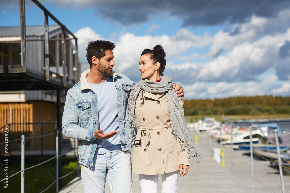 Content handsome young man in jacket walking with beloved woman and embracing her by shoulder, he sharing his story with girlfriend during stroll on pier Content handsome young man in jacket walking with beloved woman and embracing her by shoulder, he sharing his story with girlfriend during stroll on pier
