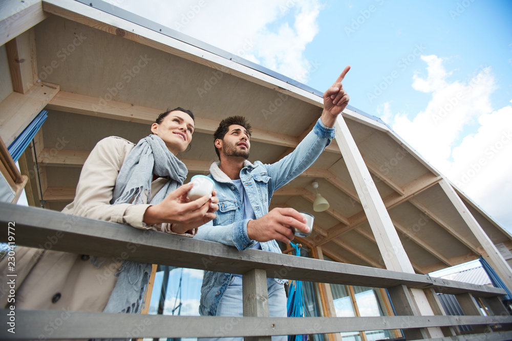 Content handsome young guy with stubble wearing denim jacket leaning on wooden railing and pointing with finger in distance while showing ship to girlfriend, they drinking tea on balcony