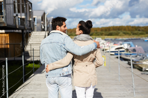 Rear view of young couple in casual clothing embracing each other and enjoying stroll on warm autumn day while walking on pier