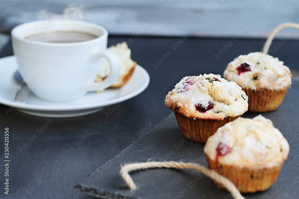 Cranberry Muffins on a slate serving tray with open muffin with butter and a steaming hot cup of coffee in the background. Extreme shallow depth of field with selective focus on center muffin. Cranberry Muffins on a slate serving tray with open muffin with butter and a steaming hot cup of coffee in the background. Extreme shallow depth of field with selective focus on center muffin.