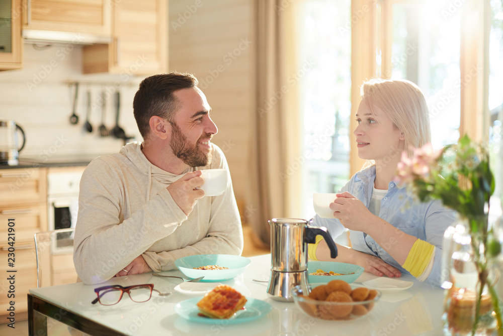 Young married Caucasian couple sitting at table in cozy kitchen, drinking coffee, eating breakfast and talking about plans for weekend Young married Caucasian couple sitting at table in cozy kitchen, drinking coffee, eating breakfast and talking about plans for weekend
