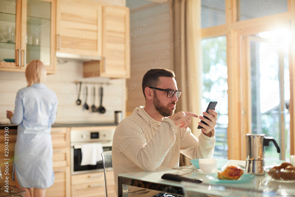 Bearded Caucasian man in glasses sitting at kitchen table and texting on smartphone while his wife washing dishes after breakfast