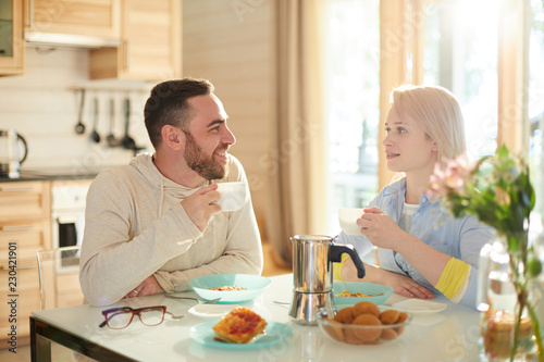 Young married Caucasian couple sitting at table in cozy kitchen  drinking coffee  eating breakfast and talking about plans for weekend