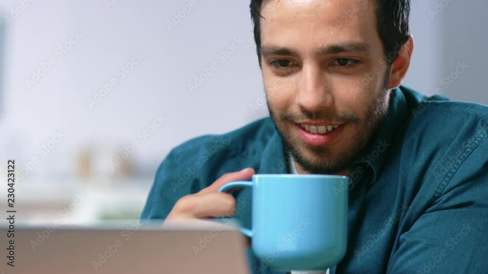 Portrait of Handsome Young Man Using Laptop Computer at Home, Watching and Laughing at Content. Happy Hispanic Man Smiles and Drinks from Mug. Portrait of Handsome Young Man Using Laptop Computer at Home, Watching and Laughing at Content. Happy Hispanic Man Smiles and Drinks from Mug.