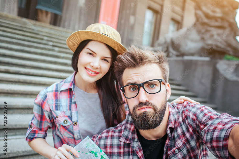 Serious bearded man holds camera and looks on it. He is upset. Nice and beautiful young woman stands behind him and smiles. She holds map.
