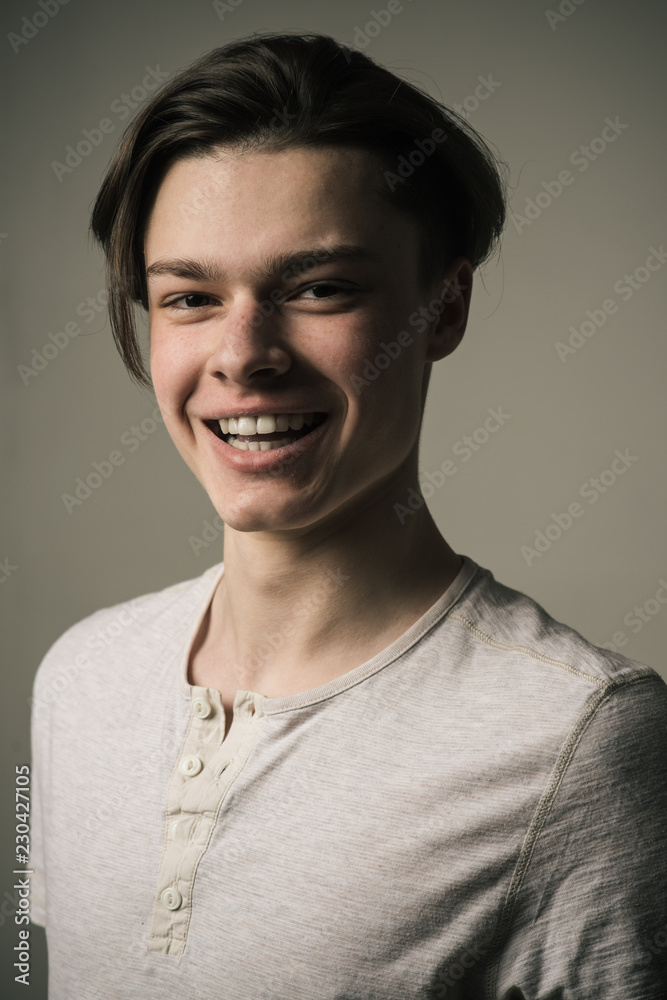 Happy smiling young man with medium length hair isolated on gray background. Cheerful teenager in white T-shirt laughing, joyful youth lifestyle concept Happy smiling young man with medium length hair isolated on gray background. Cheerful teenager in white T-shirt laughing, joyful youth lifestyle concept