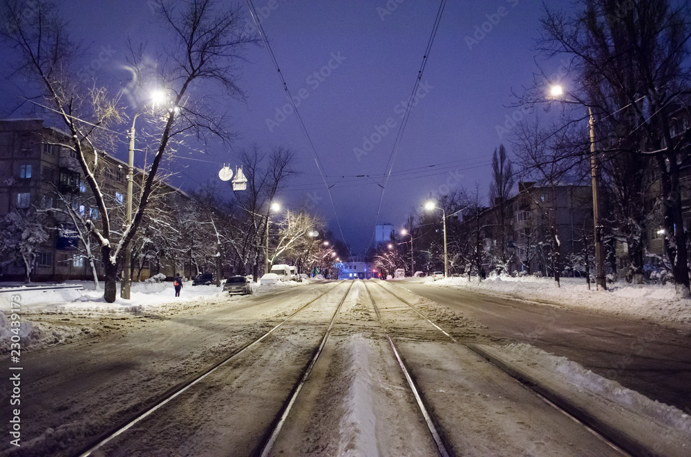 Center of the snowy road with rails. The night city with night traffic.