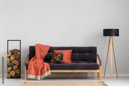 Log of wood next to comfortable sofa with strawberry red blanket and pillows, stylish wooden lamp with black lampshade, real photo copy space on the empty grey wall