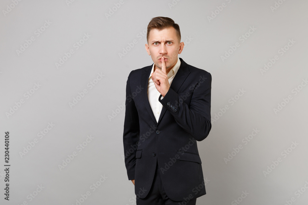 Strict young business man in classic black suit shirt saying hush be quiet with finger on lips shhh gesture isolated on grey background. Achievement career wealth business concept. Mock up copy space. Strict young business man in classic black suit shirt saying hush be quiet with finger on lips shhh gesture isolated on grey background. Achievement career wealth business concept. Mock up copy space.