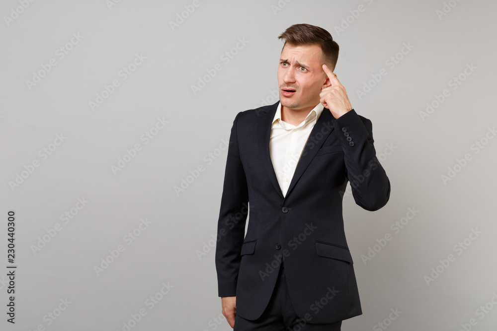Portrait of concerned young business man in classic black suit and shirt holding finger on temple isolated on grey background in studio. Achievement career wealth business concept. Mock up copy space. Portrait of concerned young business man in classic black suit and shirt holding finger on temple isolated on grey background in studio. Achievement career wealth business concept. Mock up copy space.