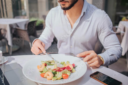 Cut view of bearded young man sitting at table and eating salad. There are laptop and phone on picture.