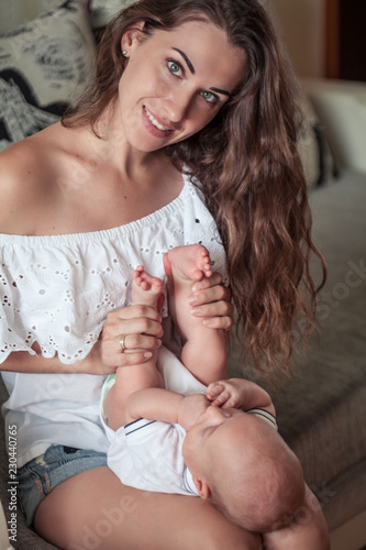 Beautiful young mother holds the baby in her arms. Mom holds the baby s legs in her hands for 3 months. Love. Motherhood. Happiness. House. Cozy.