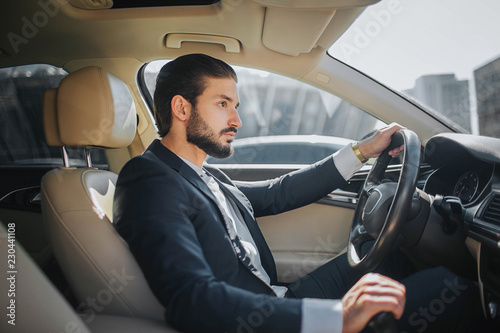 Handsome and confident young man sits in car and looks straight forward. He drives car. It looks luxury inside. He holds one hand on steering wheel and another one on handbrake.