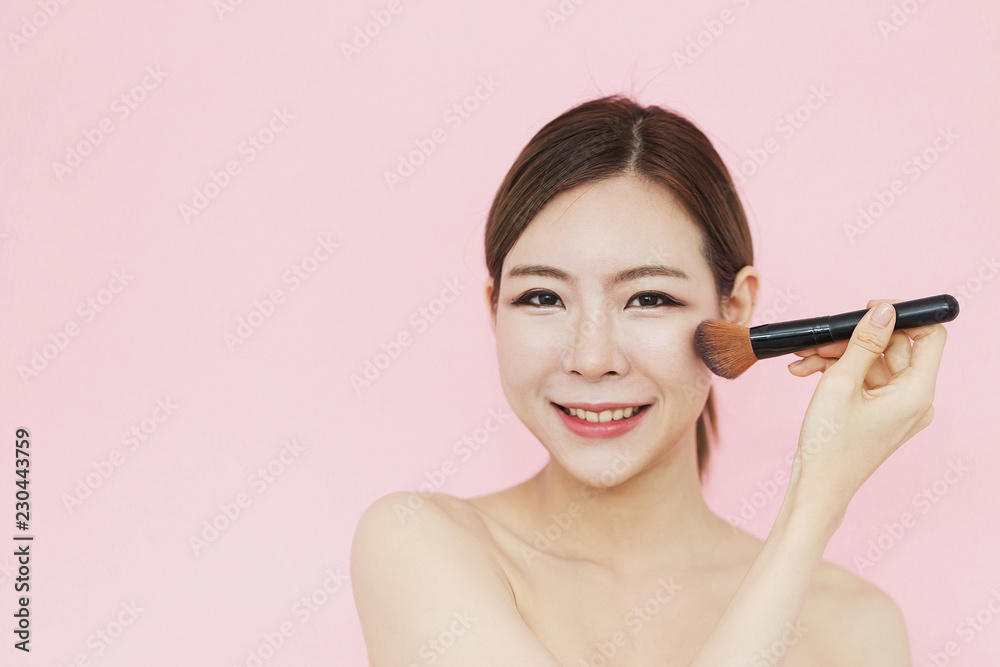 Beautiful Attractive Asian women smile with make up brush touching on her face perfect skin with copy space feeling so happiness and confident,isolated on pink background