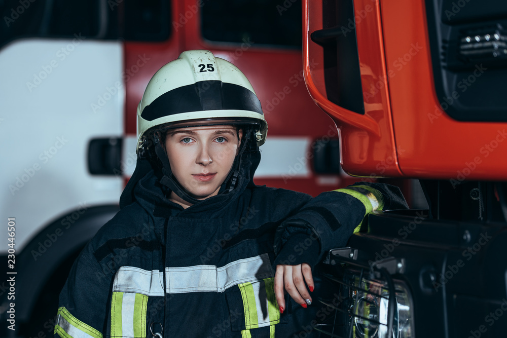Obraz premium portrait of female firefighter in helmet leaning on truck at fire station