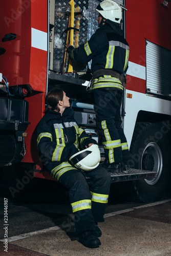 Fototapeta Naklejka Na Ścianę i Meble -  female firefighter in protective uniform looking at colleague checking equipment in truck at fire department