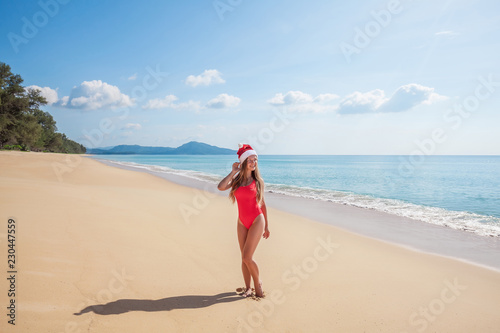 Happy young woman with long hair in red swimsuit and santa claus hat standing on the tropical paradise beach by the sea with beautiful blue water and blue sky on Phuket island,Thailand