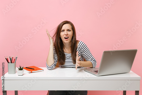 Bild auf Leinwand Young exhausted irritated woman in perplexity spreading hands sit, work at white desk with contemporary pc laptop isolated on pastel pink background