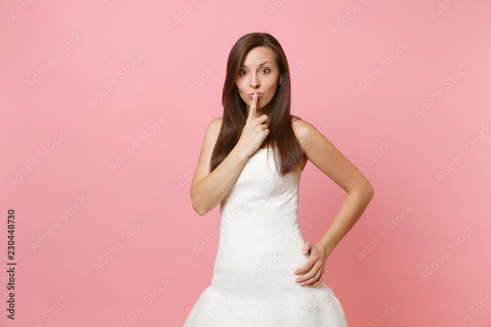 Portrait of beautiful bride woman in white lace wedding dress standing and keeping index finger on lips isolated on pink pastel background. Wedding celebration concept. Copy space for advertisement. Portrait of beautiful bride woman in white lace wedding dress standing and keeping index finger on lips isolated on pink pastel background. Wedding celebration concept. Copy space for advertisement.