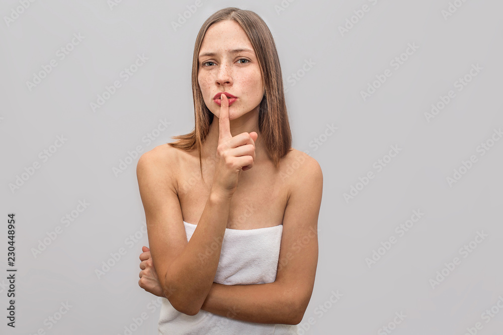 Beautiful and attractive young freckled woman stands and poses. She wears white towel. Model holds finger on red lips. Isolated on grey background. Beautiful and attractive young freckled woman stands and poses. She wears white towel. Model holds finger on red lips. Isolated on grey background.