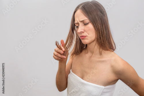 Unhappy and unsatisfied young woman stands and looks at her hair. She touches her hair and looks at it. Woman wears white towel across her body. Isolated on grey background.