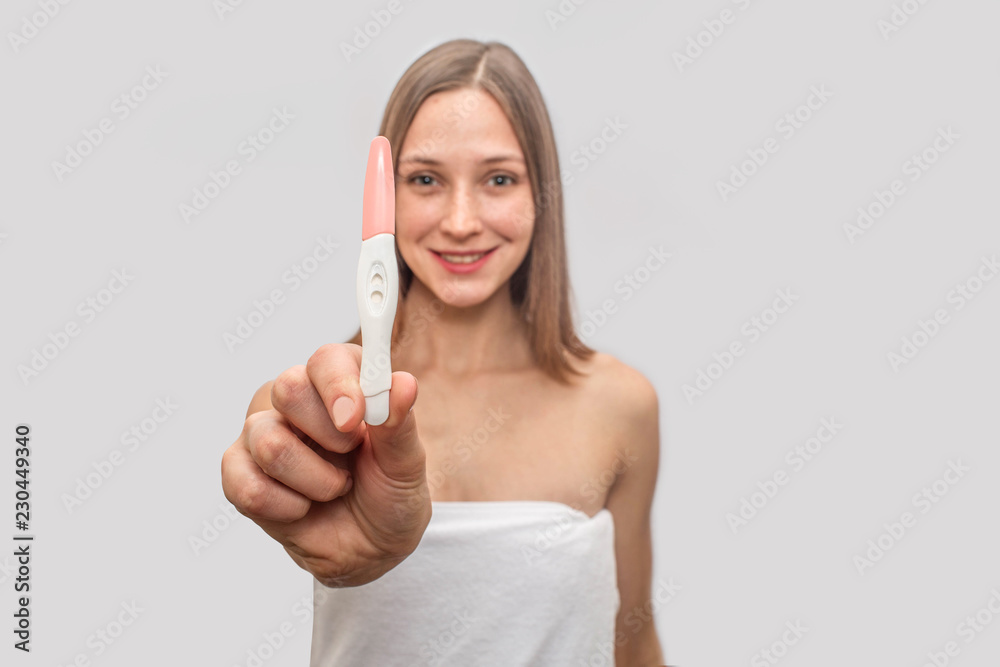 Happy young woman in white towel stands and holds pregnancy test. She is excited. Model smiles. Isolated on grey background. Happy young woman in white towel stands and holds pregnancy test. She is excited. Model smiles. Isolated on grey background.
