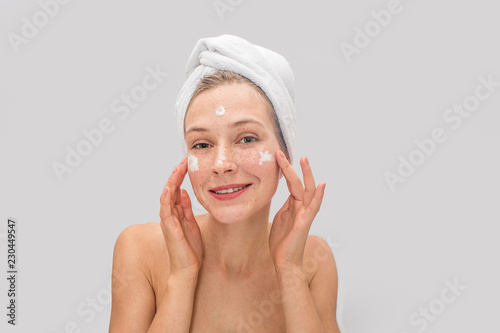 Lovely and nice freckled woman looks on camera and poses. She smiles a bit. Young woman has some cream on cheeks and forehead. There white towel on her hair. Isolated on grey background.