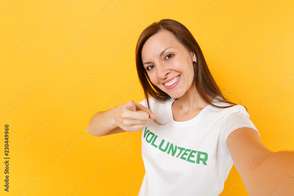 Selfie image of happy smiling satisfied woman in white t-shirt with written inscription green title volunteer isolated on yellow background. Voluntary free assistance help, charity grace work concept. Selfie image of happy smiling satisfied woman in white t-shirt with written inscription green title volunteer isolated on yellow background. Voluntary free assistance help, charity grace work concept.