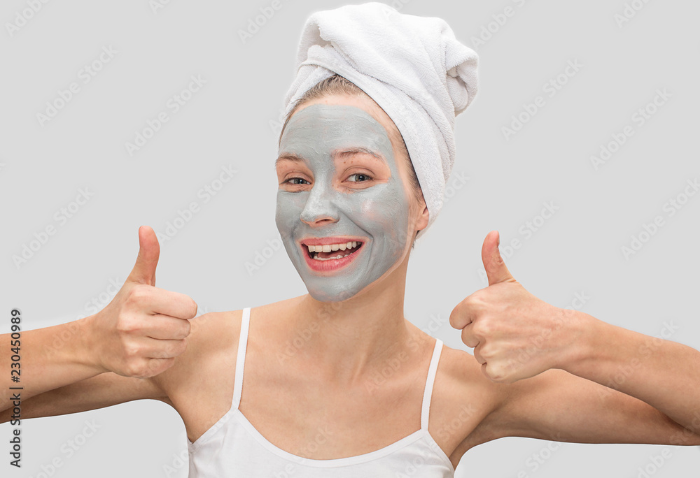 Positive and funny young woman shows her big thumbs up. Model smiles. She has mud mask on her face. Woman has white towel around her hair and t-shirt on body. Isolated on grey background. Positive and funny young woman shows her big thumbs up. Model smiles. She has mud mask on her face. Woman has white towel around her hair and t-shirt on body. Isolated on grey background.