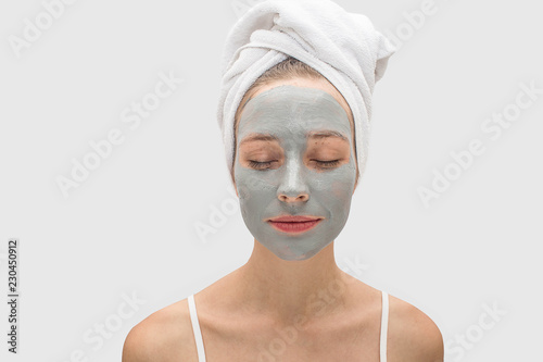 Calm and peaceful young woman stand in front of camera and keeps eyes closed. Her face is covered with mask. There are white towel on hair. Isolated on grey background.