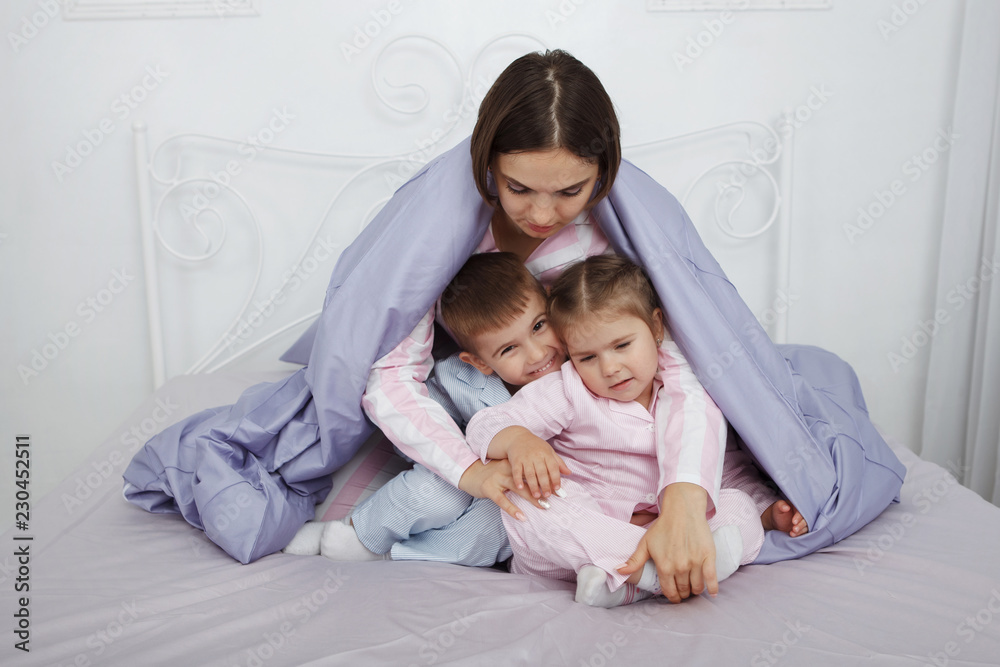 Fototapeta premium Young beautiful woman mother sitting under a blanket with children in bed with blue linens.