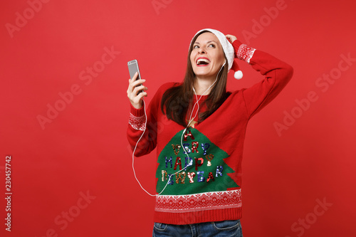 Funny young Santa girl with earphones looking up, put hand on head hold mobile phone listen music isolated on red background. Happy New Year 2019 celebration holiday party concept. Mock up copy space.