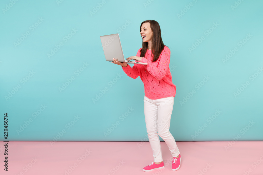 Full length portrait of woman in knitted rose sweater, white pants using laptop pc computer isolated on bright pink blue pastel wall background in studio. Fashion lifestyle concept. Mock up copy space Full length portrait of woman in knitted rose sweater, white pants using laptop pc computer isolated on bright pink blue pastel wall background in studio. Fashion lifestyle concept. Mock up copy space